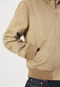 Casaco com fecho éclair em tom bege, com um poliéster ribado nas bainhas e punhos, feito de um tecido suave; possui bolsos laterais e detalhes de costura discretos.