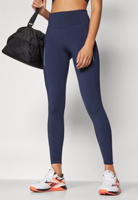 Marinehøye leggings i elastisk stoff, med en glatt tekstur, strømlinjeformet design og flatlock-sømmer, kombinert med hvite og oransje treningssko.