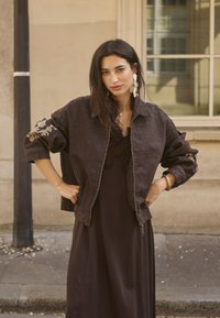 Femme aux longs cheveux foncés portant une veste marron foncé avec des manches brodées et une robe assortie, debout à l'extérieur les mains sur les hanches.