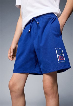Person trägt blaue Shorts mit elastischem Bund und Kordelzug, mit Tommy Hilfiger-Logo und dem Text "1985" am linken Bein.