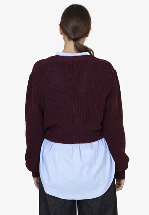 Maglione lavorato a maglia borgogna con un design corto, maniche lunghe e polsini a coste, indossato sopra una camicia azzurro chiaro con orlo curvo.