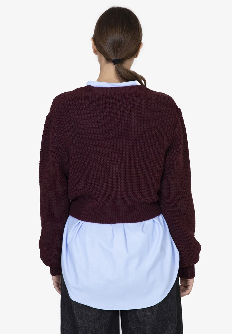 Maglione lavorato a maglia borgogna con un design corto, maniche lunghe e polsini a coste, indossato sopra una camicia azzurro chiaro con orlo curvo.