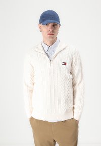 Witte kabelgebreide trui met een kwart rits. Daaronder is een lichtblauw overhemd met kraag zichtbaar. Een blauwe pet met een logo accentueert de outfit.