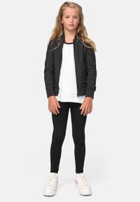 Svart bomberjakke, hvit t-skjorte, svarte leggings og hvite joggesko. Langt, bølgete hår, står med armene avslappet ved sidene, nøytral bakgrunn.