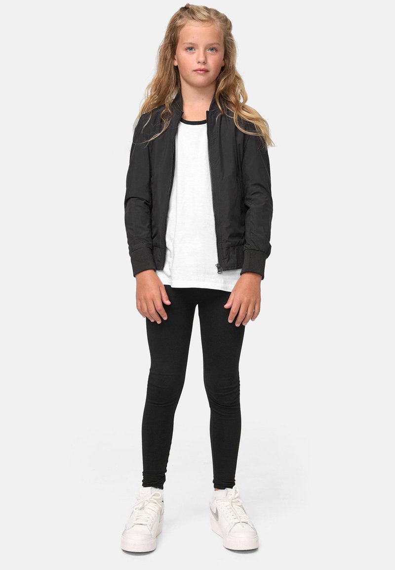 Svart bomberjakke, hvit t-skjorte, svarte leggings og hvite joggesko. Langt, bølgete hår, står med armene avslappet ved sidene, nøytral bakgrunn.