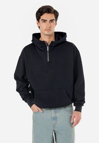 Sort zip-up hette med kengurulomme, snøring i hetten, og ribbede mansjetter, kombinert med lyseblå jeans. Myk stofftekstur.