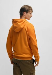 Oranje hoodie met een capuchon met trekkoord, ribbelmanchetten en een ribbelzoom. De stof lijkt zacht en wordt gedragen met olijfgroene cargobroeken.