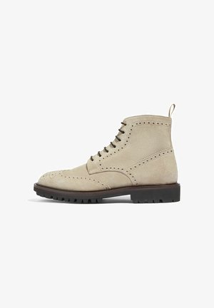 Beige ruskindsankelstøvler med brogue-detaljer, snørebåndsdesign, en løkke bagpå og en sort gummisål med struktureret greb.