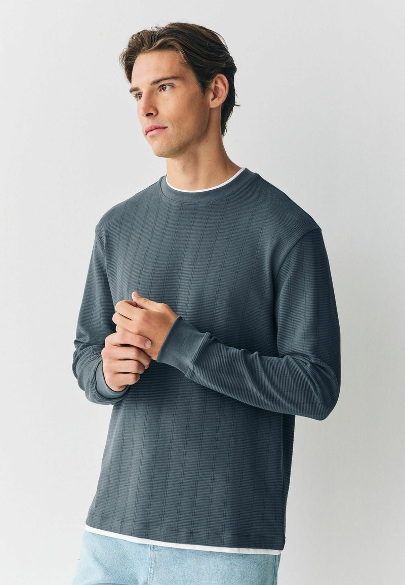 Grå langærmet skjorte med ribbet tekstur, rund hals og manchetter. Bæres over en hvid crewneck t-shirt. Casual design og pasform.
