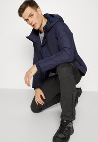 Marineblaue Kapuzenjacke mit Reißverschluss, die über einem grauen Hemd getragen wird. Schwarze, schmal geschnittene Hose und dunkle Wanderstiefel runden das Outfit ab.