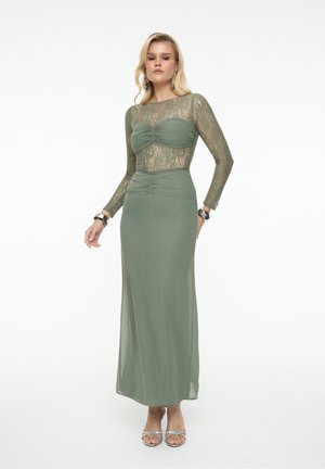 Frau in einem langen, olivgrünen Kleid mit langen Spitzenärmeln und gerafftem Oberteil, stehend vor einem schlichten weißen Hintergrund.