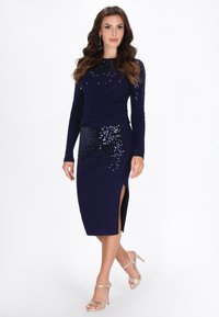Marineblaues Strickkleid mit Paillettenakzenten, langen Ärmeln und Seitenschlitz. Kombiniert mit metallischen, geschnallten Absätzen. Glatte Textur über das gesamte Kleid.