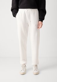 Pantaloni della tuta bianchi con coulisse, tasche laterali, gamba affusolata e texture morbida. Dettaglio del logo sulla coscia sinistra, abbinati a sneaker bianche.