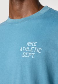 Svetlomodré bavlnené tričko s okrúhlym výstrihom a bielym potlačou na prednej strane, ktorá znie "NIKE ATHLETIC DEPT."