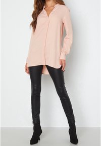 Helles rosa Blusenshirt mit V-Ausschnitt und langen Ärmeln, kombiniert mit schwarzen, kunstledernen Leggings und Ankle Boots, vor einem neutralen Hintergrund.