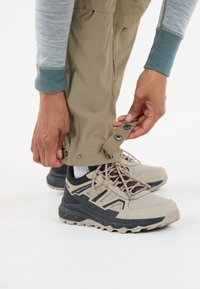 Chaussures imperméables beiges avec des accents gris et des lacets roses, associées à un pantalon marron clair en train d'être ajusté au niveau des poignets. Un haut à manches longues gris est visible.