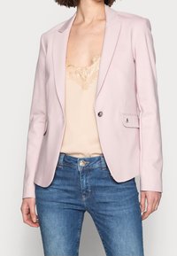 Femme portant un blazer rose clair par-dessus un haut beige orné de dentelle et un jean en denim bleu, debout devant un fond uni.