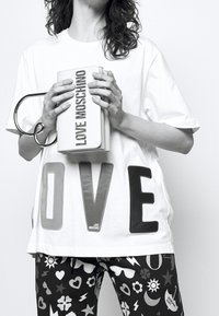 Vit t-shirt med stora "KÄRLEK"-bokstäver i grått och svart, kombinerad med svarta mönstrade byxor med vita grafiska mönster. Håller en vit clutchväska.