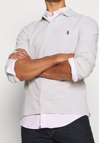 Ljusgrå långärmad polo med krage och en marinblå logotyp. Layerad över en ljusrosa och vit randig skjorta.