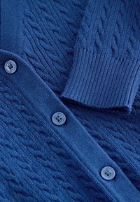 Blauer Strickcardigan mit Zopfmustern, einem gerippten Ärmelbündchen und drei blauen Knöpfen, die mit "Clarks" graviert sind, in einer Nahaufnahme.