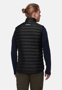 Schwarze, isolierte Weste mit gestepptem Muster und hohem Kragen, mit einem Logo auf dem Rücken, getragen über einem navyblauen Langarmshirt.