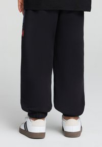 Pantaloni della tuta neri con polsini elastici, abbinati a sneakers bianche con accenti grigi e neri. Cuciture visibili e morbidezza del tessuto.