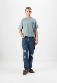 Young man standing with hands in pockets wearing a light blue graphic t-shirt, ripped blue jeans, and brown leather shoes on white background.