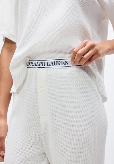 Άτομο που φοράει λευκό πουκάμισο με υφή και παντελόνι, σηκώνει το πουκάμισο για να αποκαλύψει τη ζώνη της εσωρούχου με την επιγραφή "POLO RALPH LAUREN".
