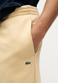 Χέρι ατόμου στην τσέπη μπεζ φόρμας με εμφανές πράσινο λογότυπο κροκόδειλου στον μηρό, φορώντας σκούρο μπλε πουκάμισο.