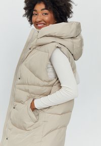 Beige puffer vest with a hood, featuring a quilted design, zip closure, side pockets, and soft texture, worn over a light long-sleeve top.