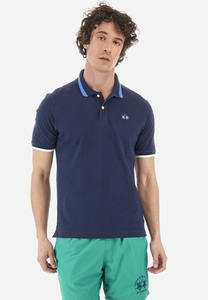 Camisa polo azul marino con un cuello azul, acentos blancos en las mangas y un logo bordado azul en el pecho. Tela de algodón suave.