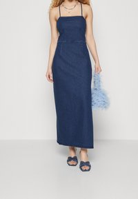 Robe maxi en denim avec des bretelles fines, un corsage ajusté et une légère évasée. Associée à un sac à main en plumes bleues et des sandales navies ornées.