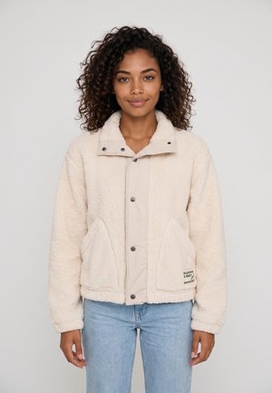 Vrouw met krullend haar die een lichtbeige fleecejas en lichtblauwe spijkerbroek draagt, staand voor een effen achtergrond.