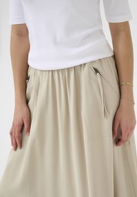 Beige sukně ke kolenům vyrobená z hladké látky, se zapínacími kapsami po stranách a elastickým pasem. Minimalistický design s jemným drapováním.