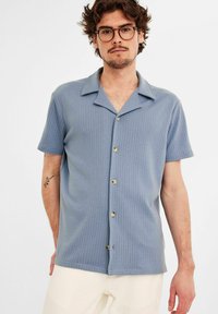 Lichtblauw shirt met korte mouwen met een gestructureerd ribbelontwerp, een gespreide kraag en koperen knopen aan de voorkant. Draag het met crèmekleurige broek.