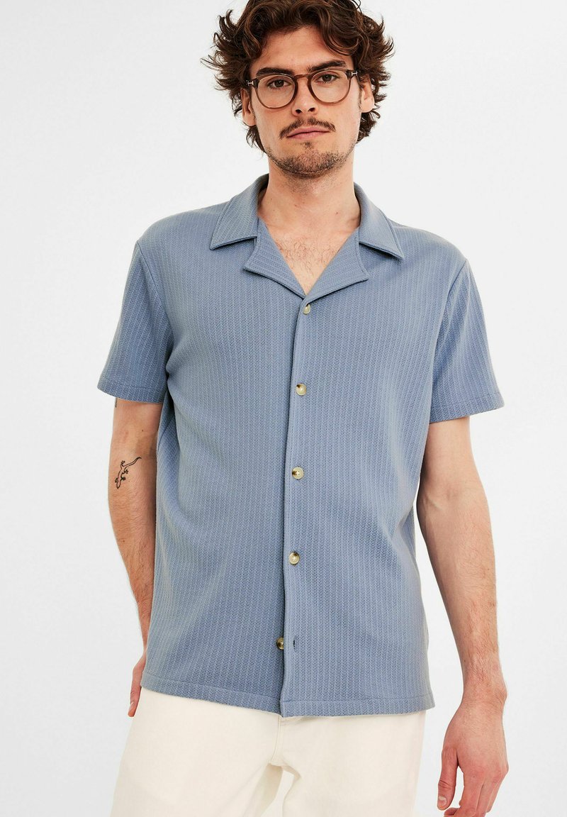 Lichtblauw shirt met korte mouwen met een gestructureerd ribbelontwerp, een gespreide kraag en koperen knopen aan de voorkant. Draag het met crèmekleurige broek.