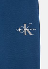 Tamsiai mėlynos medvilninės treninginės kelnes su sidabriniu logotipu, ant kurio parašyta "Calvin Klein Jeans", o didesnėmis raidėmis "CK". Lygi tekstūra, standartinė fasonas.