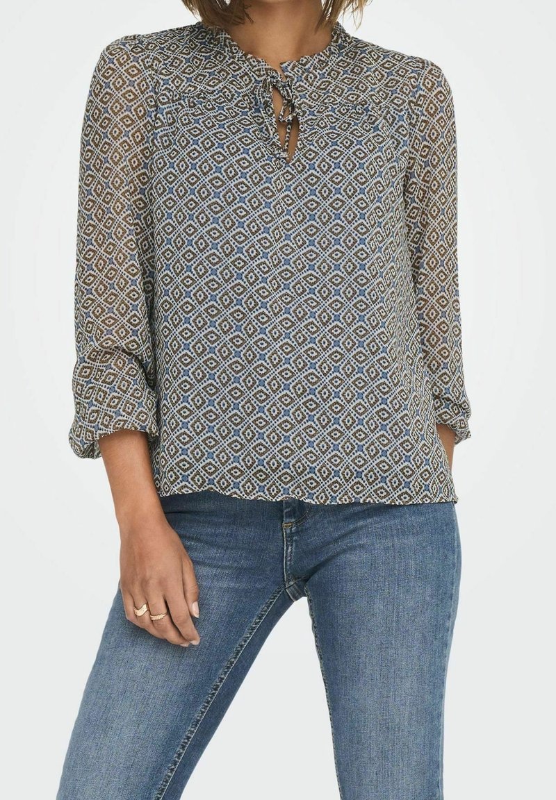 Blouse à motifs multicolores avec un décolleté en forme de trou de serrure, des épaules froncées et des manches trois-quarts, portée avec un jean bleu.