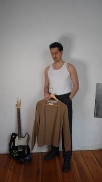Homme en débardeur blanc et pantalon noir tenant un pull marron sur un cintre, debout à côté d'une guitare électrique noire sur un sol en bois.