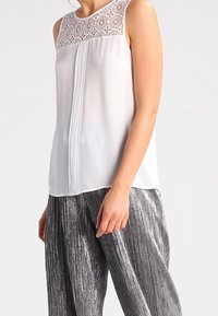 Blouse blanche sans manches avec un empiècement en dentelle, dotée d'un col en trou de serrure et de plis sur le devant, associée à un pantalon plissé métallique argent.