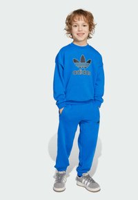 Sweatshirt Adidas bleu avec grand logo noir, associé �à un pantalon de jogging bleu assorti. L'ensemble présente une coupe décontractée et des poches latérales.