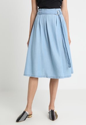 Kobieta ubrana w jasnoniebieską spódnicę midi z materiałowym paskiem i czarną bluzkę, do tego czarne, białe i brązowe wzorzyste wsuwane buty na płaskim obcasie.