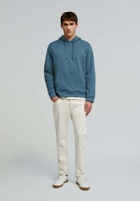 Hætte sweatshirt i blå stof, med en forlommer og justerbare snore. Matchet med lysebeige bukser og hvide sneakers.