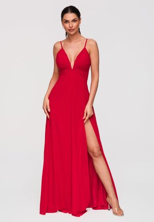 Femme portant une robe rouge sans manches avec un décolleté en V profond et une fente haute sur la cuisse, debout devant un fond clair uni.