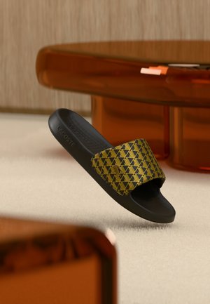 Black Lacoste slide with gold geometric pattern on strap, displayed against a beige carpet and brown translucent table background.