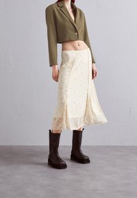 Lyhennetty oliivinvihreä bleiseri, kukkakuvioinen kerma-beige hame ja polvipituiset mustat saappaat paksuilla pohjilla. Sileä kangas, jolla on virtaava rakenne.