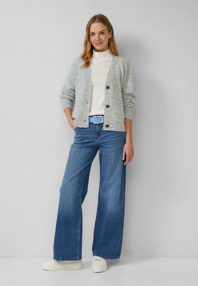 Grauer strukturierter Cardigan mit schwarzen Knöpfen, getragen über einem weißen Rollkragenpullover, kombiniert mit weit geschnittenen blauen Jeans und weißen Turnschuhen.