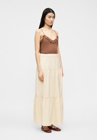 Μια γυναίκα στέκεται φορώντας καφέ αμάνικο τοπ με βολάν, κρεμ μακρύ φούστα με βολάν και καφέ δερμάτινα παπούτσια με κορδόνια, μπροστά σε ένα απλό φωτεινό φόντο.