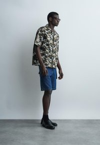 Homme portant des lunettes de soleil, une chemise camouflage à manches courtes, un short en jean, des chaussettes noires et des chaussures habillées noires, debout contre un mur clair uni.