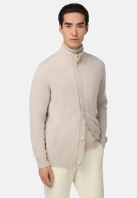 Beige stickad kofta med ribbade manschetter och krage, med fem ljusa knappar, sidofickor och en strukturerad tyg.